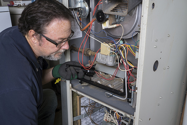 Mantoloking furnace repair technician in blue shirt wearing glasses and black and green workgloves examines an open furnace compartment with a small flashlight