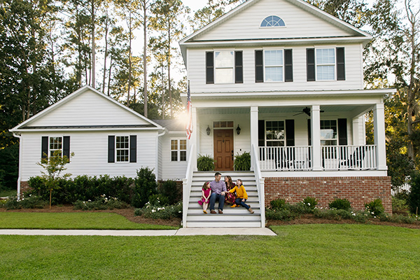 White 2-story home with black shutters and family sitting on steps leading up to porch with a green lawn in foreground